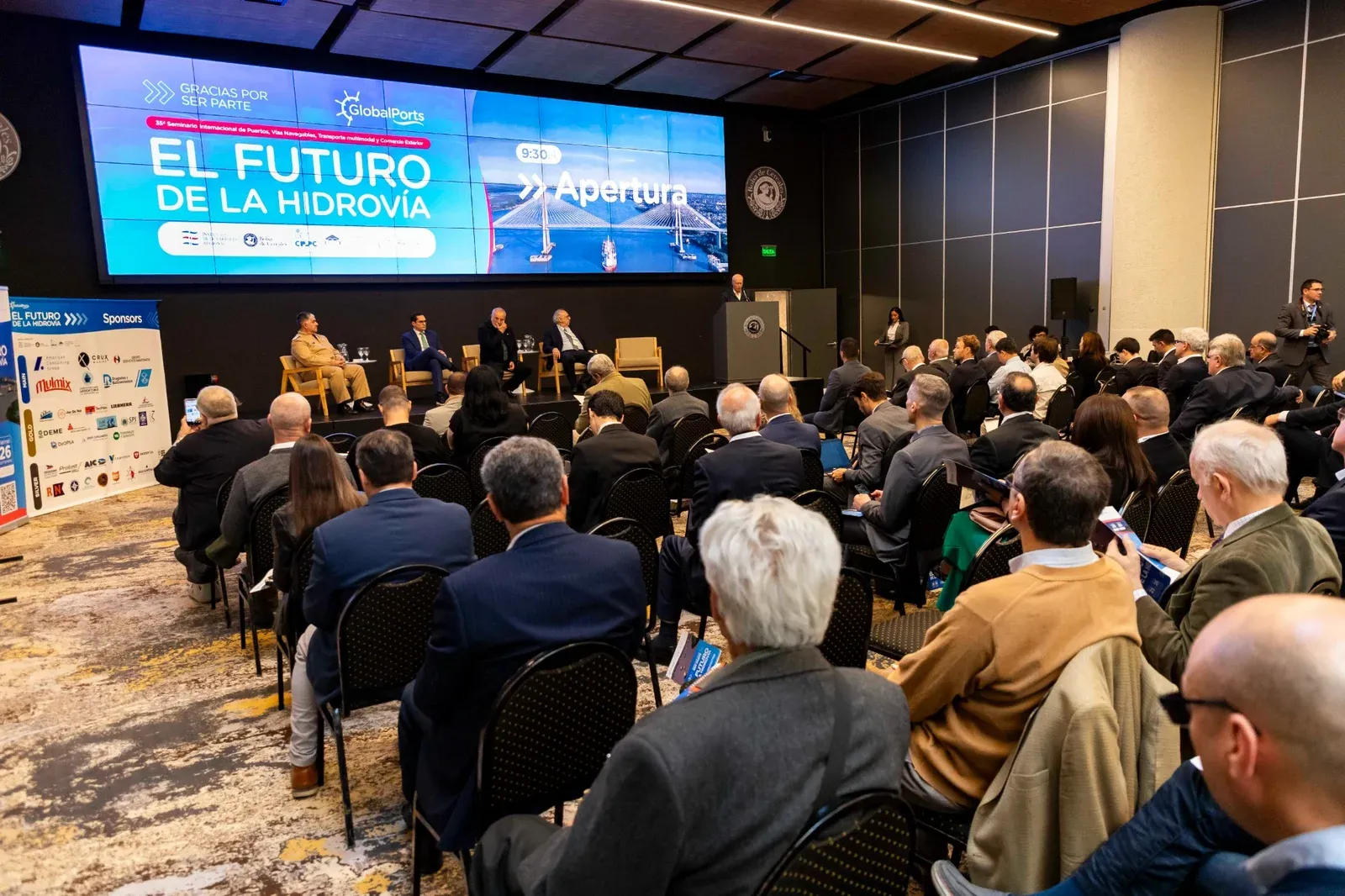 Sala llena de asistentes de espaldas frente a la pantalla del panel inaugural